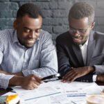 Happy African American office workers dressed in formal clothing having cheerful looks, studying and amalyzing legal documents on table using magnifying glass while getting papers ready for meeting Happy African American office workers dressed in formal clothing having cheerful looks, studying and amalyzing legal documents on table using magnifying glass while getting papers ready for meeting