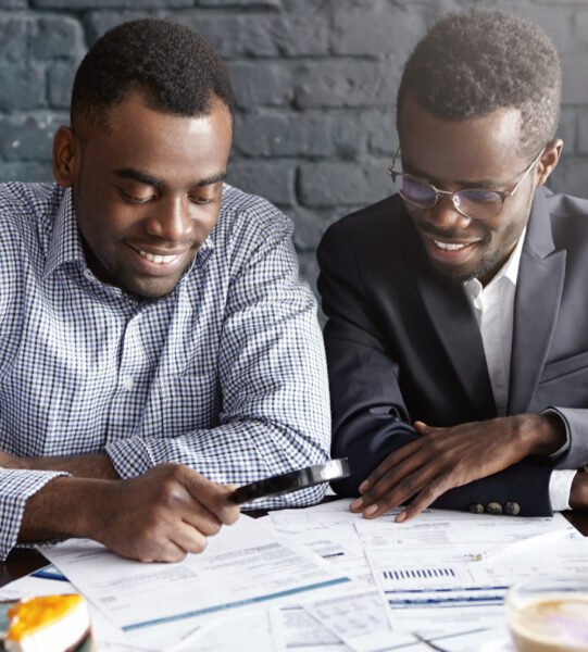 Happy African American office workers dressed in formal clothing having cheerful looks, studying and amalyzing legal documents on table using magnifying glass while getting papers ready for meeting