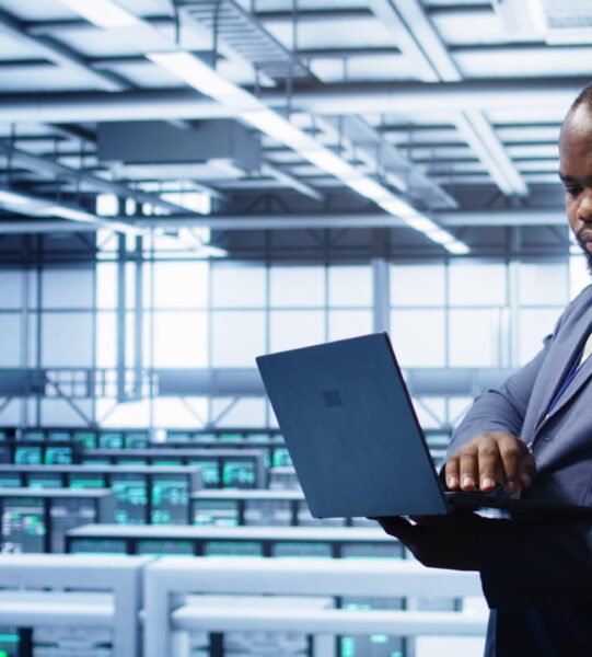 African american IT operator doing maintenance using laptop to prolong data center equipment life span. IT professional in server hub workspace monitoring server mainframes using notebook, camera B